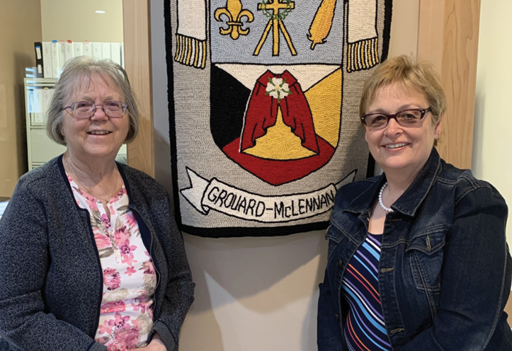 Two women standing in front of the crest of the Roman Catholic Archdiocese of Grouard-McLennan